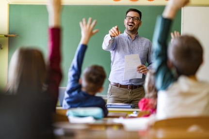 Happy male teacher aiming at his students who raised their hands to answer the question on a class at elementary school.